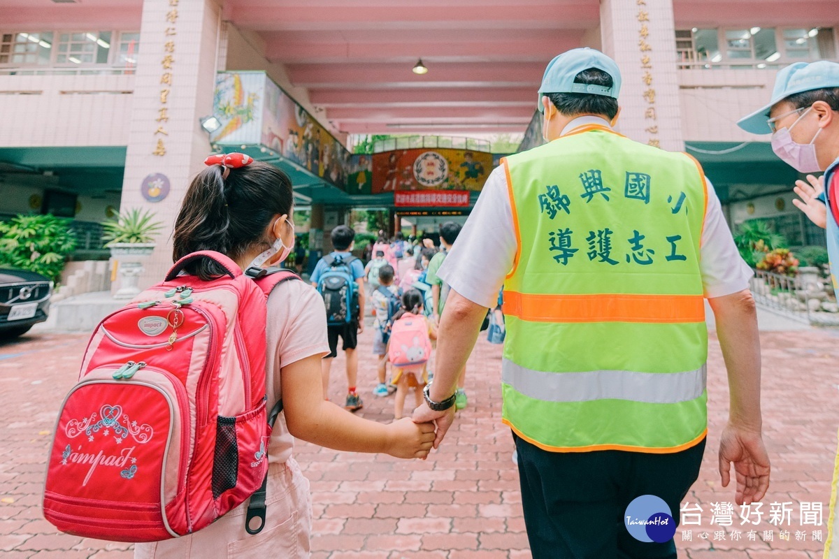 桃園市長鄭文燦穿著反光背心、手拿導護旗幟，在錦興國小校門口擔任一日導護志工，引導學童們過馬路、安全進入校園。